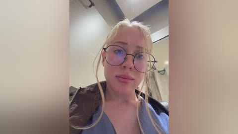 A video of a young woman with fair skin, light blonde hair, and round glasses, wearing a light blue jacket, standing in a modern, dimly lit room with beige walls.