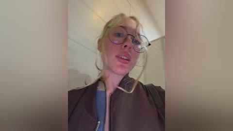 Video of a young woman with blonde hair, wearing glasses, a dark jacket, and a blue shirt, standing in an indoor corridor with white walls and a backpack strap visible.