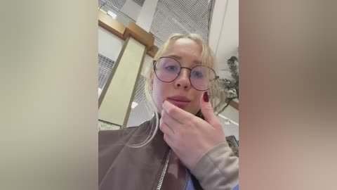 A video shows a blonde woman with glasses and a grey jacket, resting her chin on her hand, in an office with metal mesh panels and a plant.