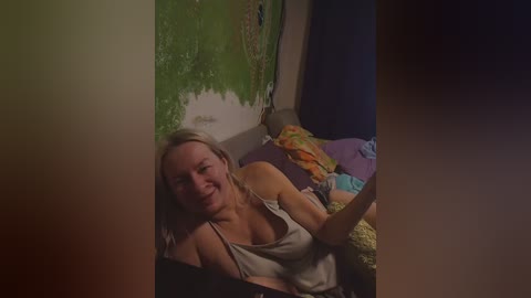 Video of a smiling, fair-skinned woman with shoulder-length blonde hair, wearing a white tank top, lying on a bed with colorful pillows and a dark curtain in the background.