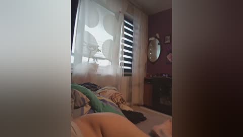 A dimly-lit video of a cluttered bedroom with a person lying on a bed covered in colorful blankets, surrounded by a messy room with pink walls and a dark wooden dresser.