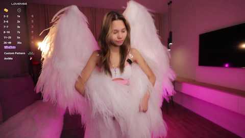 Video of a young woman with long brown hair in a white feathered angel costume, standing in a modern living room with pink and purple lighting.