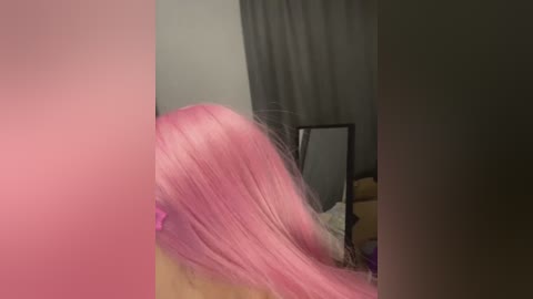 Video of a person with long, pink hair, viewed from the side, with a blurred background featuring a mirror and gray curtains.