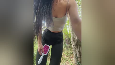 A video of a woman with long black hair, wearing a white crop top and black leggings, holding a pink hairbrush against a tree in a forest.