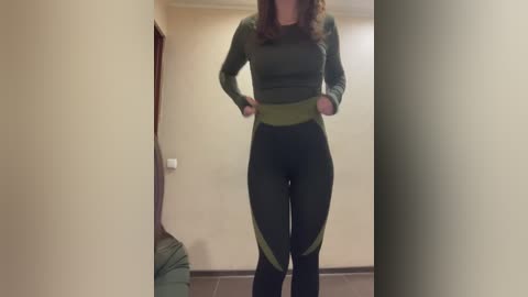 Video of a woman in a green, long-sleeve top and black leggings, standing in a beige-walled hallway with a brown couch and a wooden railing visible.