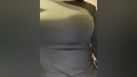 A video of a person wearing a snug, olive-green long-sleeved shirt, with a hint of a tattoo on their left arm. The background is blurred, suggesting an indoor setting.