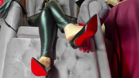 Video of a woman wearing tight, shiny black leggings, red-soled stilettos, and a red top, reclining on a plush, grey, tufted sofa, with a person in a red shirt and black jacket in the background.