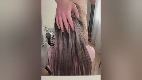 A video shows a woman with long, straight, dark brown hair being massaged by a man's hand. She is seated, wearing a pink top. The background features a zebra-patterned pillow and a curtain.