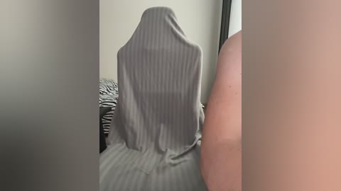 Video of a person sitting in a chair covered by a grey, striped blanket, revealing only their back and part of their arm, in a dimly lit room with a zebra-patterned bedspread visible in the background.
