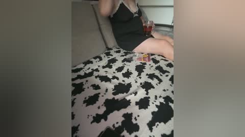 Video of a person in a black, lace-trimmed nightgown, lounging on a bed with a black-and-white cow patterned blanket. The background shows a neutral-colored room with a glass of red wine on a bedside table.