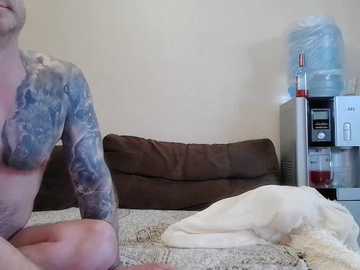 Video of a shirtless man with a large, colorful tattoo on his right arm, sitting on a bed with beige sheets, brown couch behind him, water dispenser on the right, and a white towel on the bed.