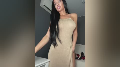 Video of a slender woman with long black hair, wearing a beige, sleeveless dress, standing in a modern, minimalist room with gray walls and a white table.