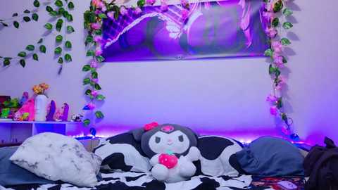 A vibrant, cozy room with a plush bed adorned with a black and white striped blanket, a pink stuffed animal, and a plush bear. The walls are decorated with pink and green hanging plants, and a large TV screen shows a dancing anime character.