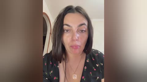 Video of a light-skinned woman with straight dark hair, wearing a black floral dress, and a gold pendant necklace. She has a neutral expression and is indoors with a wooden door and white walls in the background.