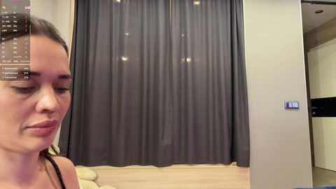 Video of a woman with light skin and dark hair in a grey tank top, leaning against a closed door with a grey curtain. Background shows a modern interior with wooden flooring.