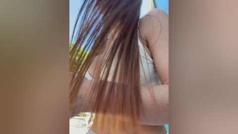 A close-up video of a woman's long, straight brown hair, captured in sunlight, partially obscuring her face. She wears a light-colored top, suggesting a relaxed, outdoor setting.