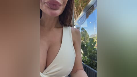 Video of a woman with light skin and brown hair, wearing a low-cut white dress, outdoors with lush greenery and a clear blue sky visible in the background.