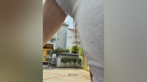 Video of a person's lower torso in white pants, captured from a low angle, showing their legs and buttocks. Background features a sunlit residential area with a yellow truck and multiple-story buildings.