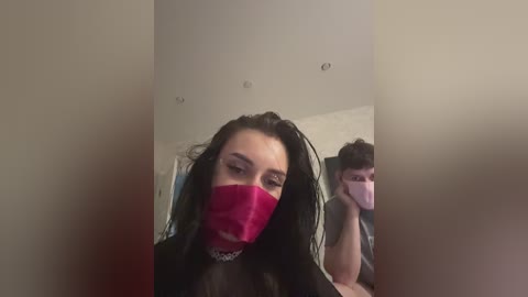 A video of a young woman with long, dark hair, wearing a pink face mask and black clothing, taking a selfie in a dimly lit room with a mirror reflection of a man in the background.