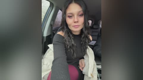 Video of a young woman with long, wavy black hair and light skin, wearing a black turtleneck, maroon pants, and a white jacket. She sits in the backseat of a car, looking contemplative.