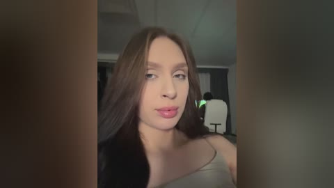 Video of a Caucasian woman with long brown hair, light skin, and pink lipstick, wearing a gray tank top, seen from the waist up, indoors with dim lighting, partially obscured by a vertical frame.