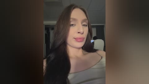 Video of a young woman with long, straight black hair and fair skin, wearing a white off-shoulder top, standing in an indoor setting with dim lighting.