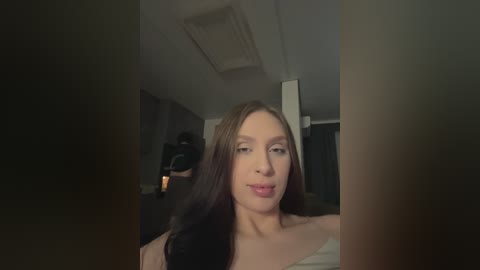 A video of a young woman with long, dark brown hair and fair skin, wearing a white top, standing indoors with dim lighting.