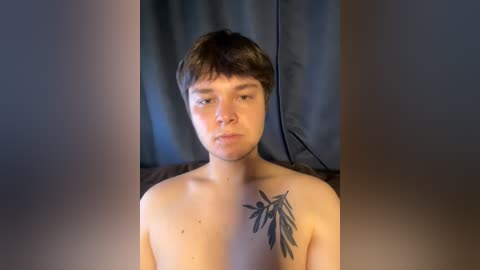 Video of a young, light-skinned, shirtless man with short brown hair, featuring a black leaf tattoo on his right shoulder, standing against a dark curtain background.