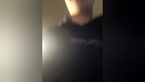 Video of a blurred person with light skin and short hair, wearing a black shirt, standing in a dimly lit room. The background is dark and indistinct.