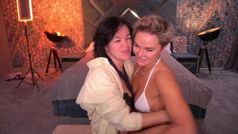 Video of two women, one in a white bikini, hugging intimately in a dimly lit, modern bedroom with stone walls, a bed, and a spotlight.