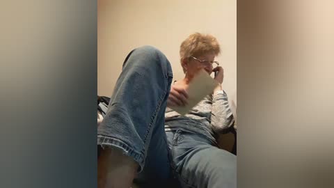 Video of an elderly woman with short, curly hair, wearing glasses and a light grey shirt, sitting on a couch with her legs up, holding a phone, against a beige wall.