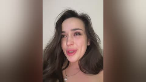 Video of a smiling woman with long, wavy dark hair, fair skin, and subtle makeup, wearing a silver necklace. Background is blurred, creating a soft focus effect.