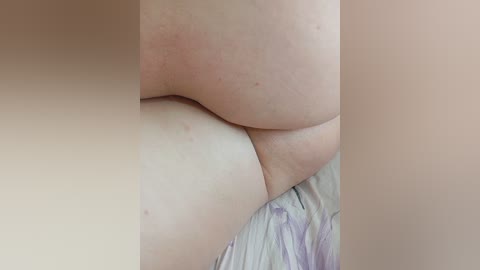 A close-up video of two nude people lying together, focusing on their buttocks and thighs. The skin is pale and smooth, with visible pores and slight redness. The background shows a bed with floral-patterned sheets.