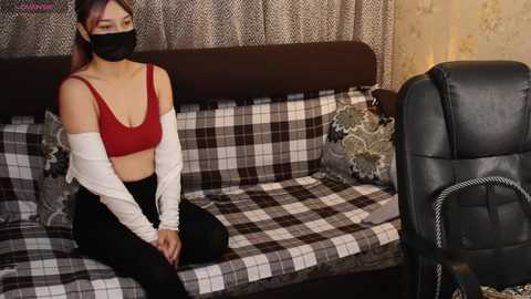 A video of an Asian woman with light skin, wearing a black face mask, red crop top, black pants, and white jacket, sitting on a checkered couch in a dimly lit room.