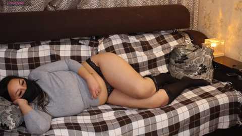 Video of a woman with medium skin tone and black hair, lying on a plaid-covered couch, wearing a grey top and black underwear, in a cozy room with a lit lamp.