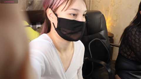 Video of an Asian woman in a white top and black face mask sitting in a black office chair. Background features a beige wall with floral patterns and a lamp.