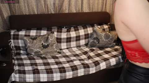 Video of a woman in a red lace bra standing next to a plaid-covered sofa with two decorative pillows, in a cozy room with patterned curtains.