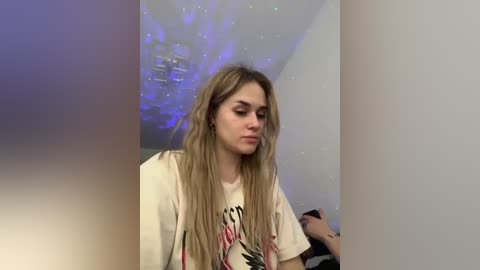 Video of a young woman with long, wavy blonde hair, wearing a graphic T-shirt, standing in a dimly lit room with a blue LED light on the ceiling.