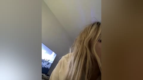 A video of a blonde woman in profile, leaning against a wall with a beige shirt, watching a TV displaying a snowy mountain landscape.