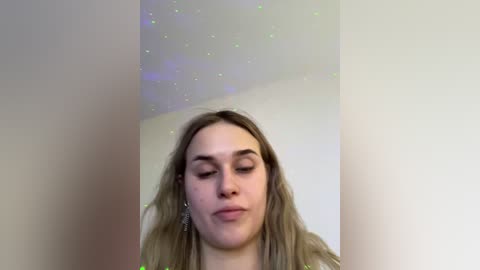 Video of a young woman with light skin, long blonde hair, wearing a light top, standing against a beige wall with a faint, sparkling pattern.