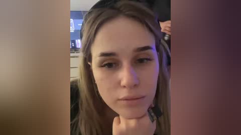 Video of a young woman with fair skin and straight brown hair, looking contemplative, indoors, with blurred background.