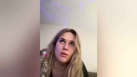 A video of a young woman with long, blonde hair, pale skin, and light makeup, sitting indoors with a plastic bag in the background, looking up thoughtfully.