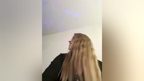 Video of a young woman with long, straight, blonde hair, wearing a black jacket, standing against a plain, white wall with faint purple reflections.