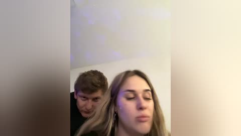 Video of a young couple in a dimly lit room. The man, with short brown hair, is slightly behind the woman, who has long blonde hair. Both have a neutral expression. The background is plain white.