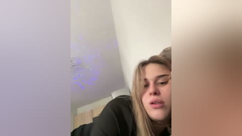 Video of a young woman with light skin and straight, shoulder-length blonde hair, partially obscured by a large, blurred object. She has a neutral expression. The background features a white ceiling with a faint, purple-hued light fixture.