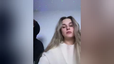 A video of a young woman with long, wavy blonde hair, wearing a white sweater, standing against a softly lit, starry backdrop. The image is slightly blurred, with a dark figure partially visible on the left.