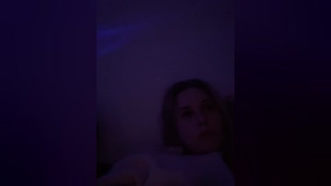 A dimly lit video shows a woman with shoulder-length brown hair, partially obscured by shadows, in a dark room. She has a neutral expression and is wearing a white top.