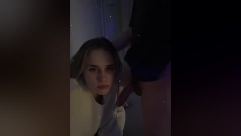 Video: A dimly lit, close-up of a blonde woman with a serious expression, kneeling in front of a dark-skinned woman with a large black purse.