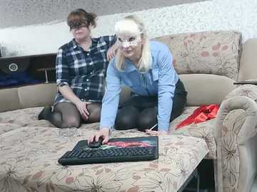Video of two women in a living room; one in a blue shirt, black skirt, and glasses, another in a plaid shirt, both kneeling on a floral-patterned sofa, typing on a laptop.