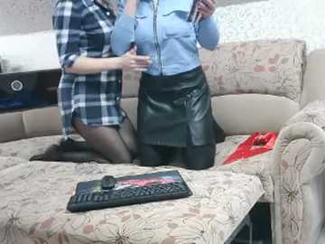 A video depicts a woman in a blue shirt and black leather skirt, kneeling on a beige floral-patterned couch, holding a smartphone. A black keyboard and red clothing lie on the couch.